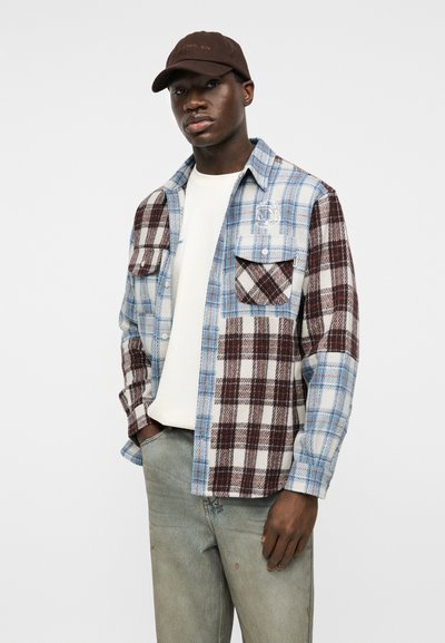 Mladý muž s hnědou čepicí, bílou košilí a modro-vínovou kostkovanou košilí s kapsami, stojící s rukama v kapsách.