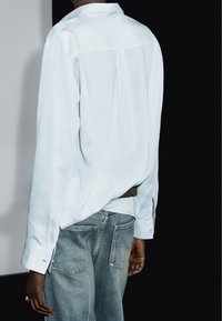 Light blue long-sleeve shirt with a back pleat, paired with faded blue jeans. The shirt features buttoned cuffs and a small logo detail.