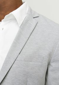 Light grey blazer with a smooth texture, notch lapels, and a chest pocket, worn over a white collared shirt. Subtle stitching detail visible.