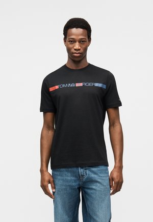 Νέος άνδρας που φοράει μαύρο T-shirt Tommy Hilfiger με κόκκινο και μπλε λογότυπο, σε συνδυασμό με μπλε τζιν, στέκεται μπροστά σε απλό φωτεινό φόντο.