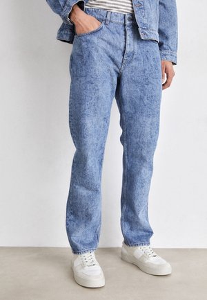 Persoon draagt lichtblauwe acid-wash jeans, witte sneakers, een gestreept shirt en een bijpassend spijkerjack, met één hand in de zak.