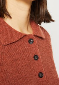 Trui in roestkleurig, gestructureerd gebreid materiaal met een kraag en drie zwarte knopen aan de voorkant. Close-up weergave die de stof en details benadrukt.