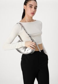Sac à main en métal argenté avec une forme incurvée, doté d'une bandoulière longue et d'accents cloutés. Présenté sur un fond neutre et associé à un haut ajusté.