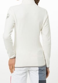 Mujer vistiendo una chaqueta de forro polar blanca con un parche en la manga, combinada con pantalones bloqueados en blanco y gris, vista desde atrás.