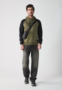 Sweat-shirt vert olive avec des manches noires, poche kangourou et cordons; associé à un jean noir délavé et des baskets noires.