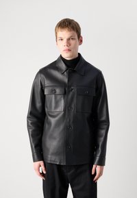 Chaqueta de cuero negra con dos bolsillos en el pecho, cierre de botones, cuello puntiagudo y mangas largas. Textura suave, diseño a medida.