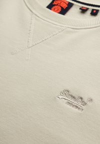 Sudadera color beige claro con una tela texturizada, que presenta un detalle de costura en forma de V en el cuello y un sutil logo bordado en el pecho.