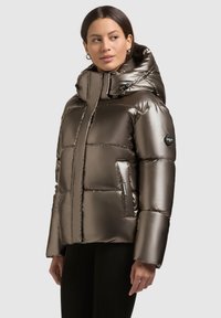 Metallic brun pufjakke med hætte, der har en lynlåslukning, store kviltede sektioner og sidelommer med et logo-patch på ærmet.