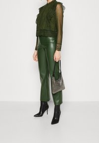 Top verde in pizzo con scollatura arricciata e maniche lunghe, abbinato a pantaloni in ecopelle verde lucida. Stivaletti neri con punta a punta e borsa in rete.