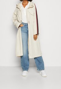 Béžový dlouhý kabát s kapucí, stahovací pas, dvě přední kapsy a vínové postranní pruhy. Nosený přes bílé tričko a modré široké kalhoty.