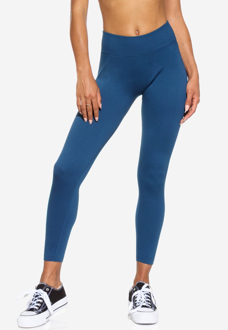 Leggings taille haute bleus en tissu lisse et élastique, associés à des baskets en toile noires avec semelles et lacets blancs.