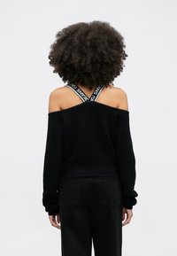 Person med lockigt hår som bär svart off-shoulder tröja som avslöjar korslagda märkta band, kombinerat med svarta byxor, stående mot vit bakgrund.