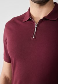 Burgunderfarget polo-skjorte med glidelås foran, myk tekstur og kortermet. Stoffet har en jevn, tett passform og minimal detaljering.