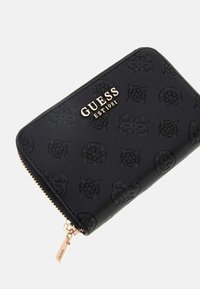Melnā jostas maku ar reljefām rakstām, zelta logotipu "GUESS EST1981" un zelta tonēta rāvējslēdzēja vilkšanas detaļu.