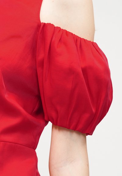 Κόκκινο μανίκι με φουσκάλες από λείο ύφασμα σε γυμνό χέρι, με ελαστική ρίγα μαζεμένη στον ώμο και το τελείωμα, σε απλό ανοιχτό φόντο.