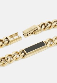 Goldkette Armband mit großen Gliedern, einem schwarzen rechteckigen Akzent und einem Verschlussmechanismus für sicheren Halt. Eingraviertes Logo sichtbar.