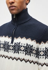 Sweter dziany z wysokim kołnierzem, granatową prążkowaną fakturą i kremowym korpusem ozdobionym wzorami śnieżynek i zygzaków w kolorach granatowym i brązowym.