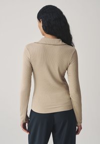 Top beige claro de manga larga con textura acanalada y cuello doblado, combinado con pantalones oscuros. Vista trasera que muestra una silueta ajustada.