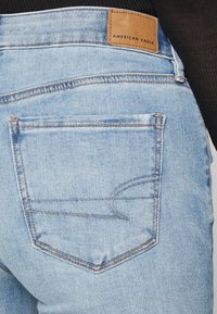 Ανοιχτό μπλε τζιν παντελόνι με σχέδιο πίσω τσέπης, που φέρει ένα καφέ δερμάτινο παpatch με την επιγραφή "AMERICAN EAGLE." Η υφή είναι λεία και ελαφρώς φθαρμένη.