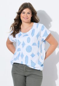 Camiseta blanca de manga corta con patrones geométricos azules, que presenta rayas y diferentes formas, combinada con pantalones verde oliva.