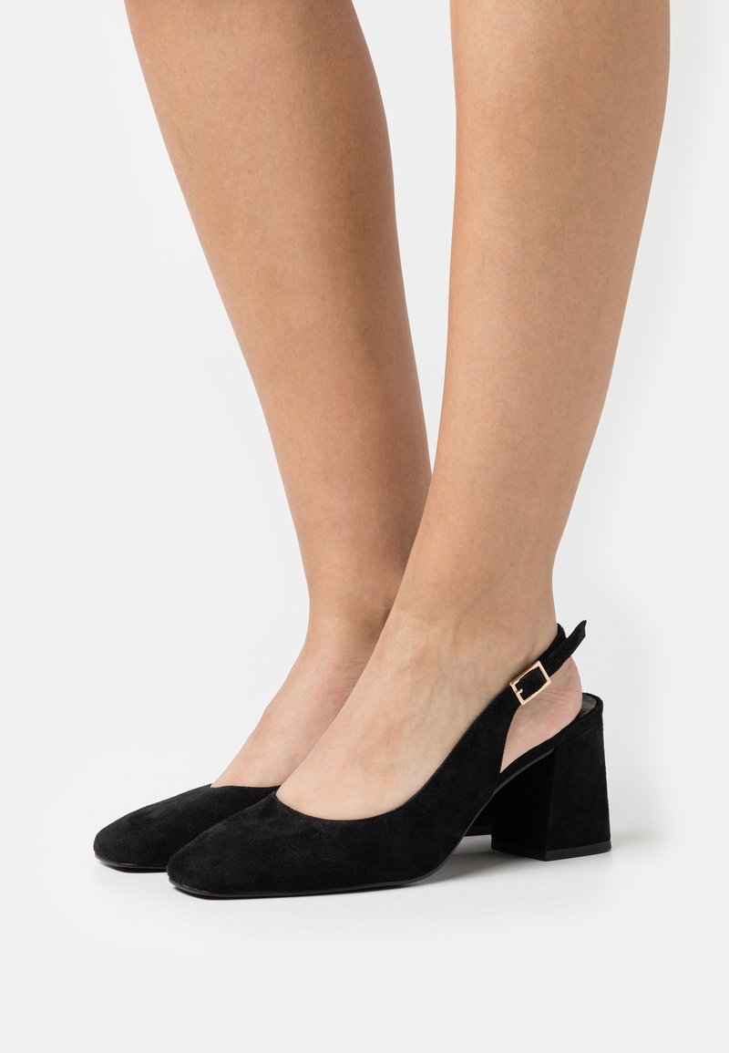 Jambes portant des talons blocs slingback en daim noir avec boucles dorées, debout sur une surface blanche.