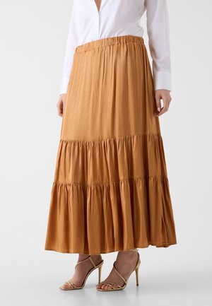 Frau trägt einen mehrstufigen, hellbraunen Maxirock mit elastischem Taillenbund, kombiniert mit einem weißen Langarmshirt und goldenen High-Heel-Sandalen.