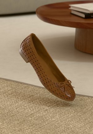 Braune, durchbrochene Ballerinas mit runder Zehenpartie, kleinem Schleifendetail und niedrigem Absatz, die über einem strukturierten Teppich liegt.