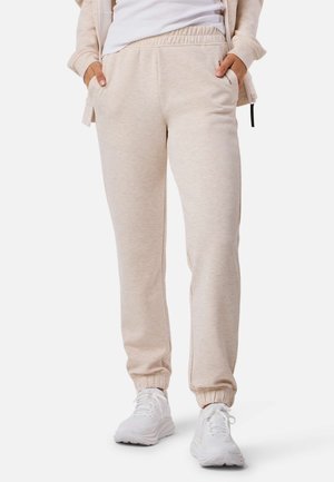 Osoba ubrana w beżowe spodnie dresowe z elastycznymi mankietami, białe trampki, biały top oraz jasnobeżową kurtkę z rękami w kieszeniach.