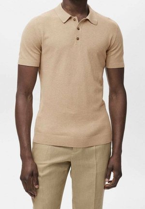 Mand iført en beige kortærmet poloshirt med tre knapper og matchende beige bukser, stående mod en ensartet lys baggrund.