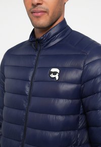 Granatowa kurtka pikowana o poziomym, przeszywanym wzorze, z wysokim kołnierzem, przednim zamkiem błyskawicznym oraz małą naszywką z logo na piersi.