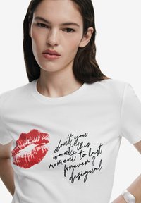 T-shirt en coton blanc avec un imprimé de rouge à lèvres rouge et un texte en cursive noir qui dit : "ne veux-tu pas que ce moment dure pour toujours ?"