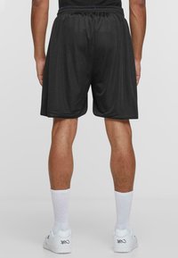 Sorte mesh treningsshorts med elastisk strikk i livet, med en løs passform og kne-lengde; brukt med hvite sokker og joggesko.