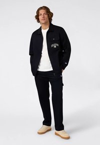 Veste noire avec un logo sur la poitrine, portée sur une chemise blanche et un pantalon noir. La tenue a un style décontracté avec des chaussures beiges.