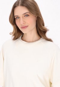 Tshirt oversize color crema con scollatura rotonda e maniche raglan corte, caratterizzato da un sottile logo dell'ancora sul lato sinistro del petto. Tessuto morbido.