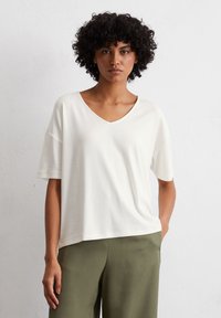 White short-sleeve t-shirt with a V-neck, made from soft fabric. Features a relaxed fit and minimal design, paired with olive green pants.