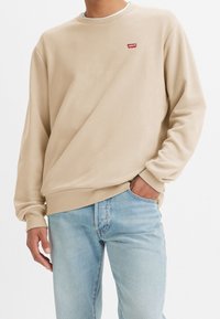 Beige sweatshirt med ribbad rund hals, långa ärmar och en liten röd Levi's logotyppatch på bröstet. Matchat med ljusblå denimjeans.
