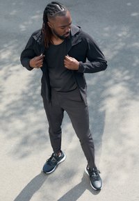 Schwarzes Sportoutfit, das aus einer leichten Jacke und enganliegenden Hosen besteht, beide aus glattem Stoff, kombiniert mit schwarzen Laufschuhen.