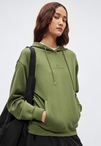 Olivovo zelená mikina s kapucňou na šnúrku, predným vreckom a jemným logom. Charakterizovaná je mäkkou textúrou a pohodlným strihom, doplnená o čiernu tašku.