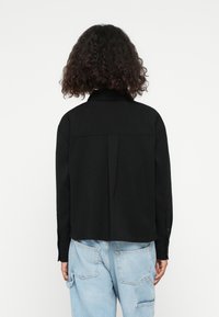 Black, cropped button-up shirt with a collar, long sleeves, and a central pleat at the back; worn over light blue baggy jeans.
