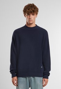 Maglione a coste di colore blu navy con scollatura a girocollo e maniche lunghe, dotato di un piccolo logo sull'orlo, abbinato a jeans azzurri.