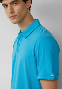 Turquoise poloshirt met een knoopsluiting bij de kraag en opgerolde mouwen. Gemaakt van een gladde stof, met een enkele knoop bij de manchetten.