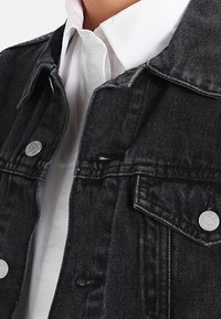 Czarna kurtka dżinsowa z srebrnymi zatrzaskami i klasycznym kołnierzem, noszona na białej koszuli. Materiał jest lekko postarzany.