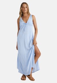 Lichtblauwe maxi-jurk met een V-hals, strikdetail bij de buste en een zijsplit, gemaakt van zachte stof met een subtiel patroon. Bruine sandalen.
