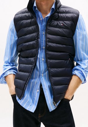 Uomo che indossa un gilet nero trapuntato senza maniche sopra una camicia a righe blu con maniche arrotolate e pantaloni scuri, mani nelle tasche.
