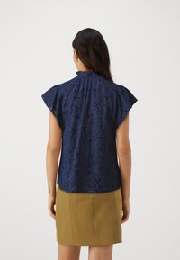 Blusa senza maniche blu navy con motivi floreali texture, colletto a volant e maniche a farfalla, abbinata a una gonna color tan.