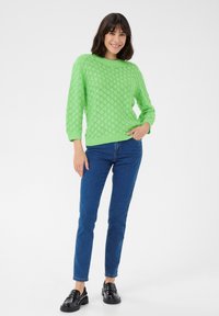 Pull en tricot vert avec un motif en losanges, encolure ronde et manches trois-quarts, associé à un jean bleu et des mocassins noirs.
