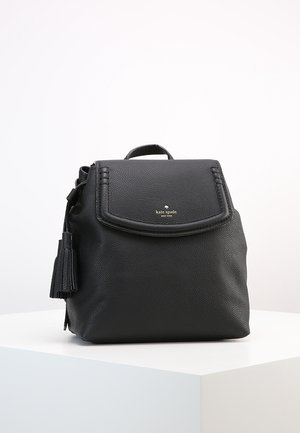 Petit sac à dos en cuir noir avec détail pompon et logo "kate spade new york" sur le rabat avant, posé sur une surface blanche.