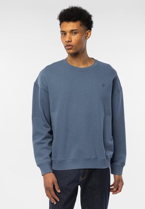 Ung mann iført en løs blå crewneck-sweatshirt og mørke jeans står foran en ensfarget hvit bakgrunn.