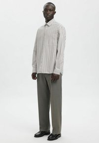 Stripet skjorte med knapper i lys beige og brun, kombinert med skreddersydde, grå bukser og svarte sko. Har brystlomme og lange ermer.