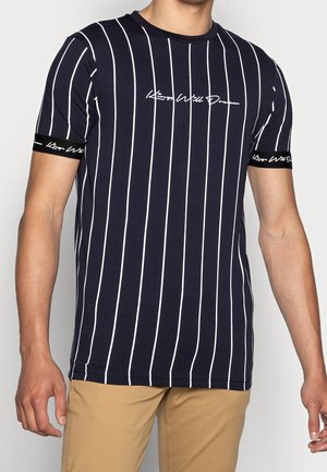 Mann trägt ein marineblaues T-Shirt mit vertikalen weißen Streifen und weißem Schriftzug auf der Brust und an den Ärmelbündchen, kombiniert mit einer beigefarbenen Hose.